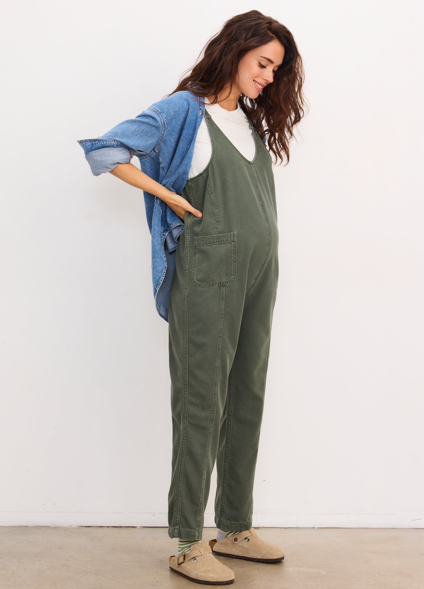 Olive green maternity jumpsuit with loose fit and tapered legs, worn over a white T-shirt and paired with a blue denim shirt