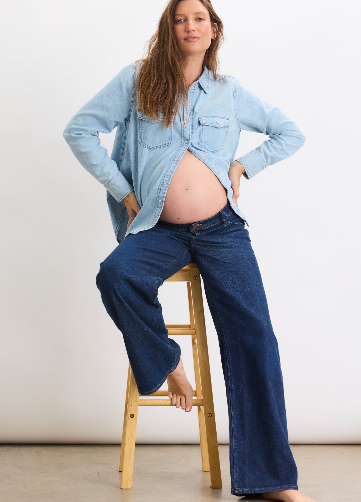 Light blue denim maternity shirt paired with dark blue relaxed wide-leg maternity jeans, barefoot model seated on wooden.