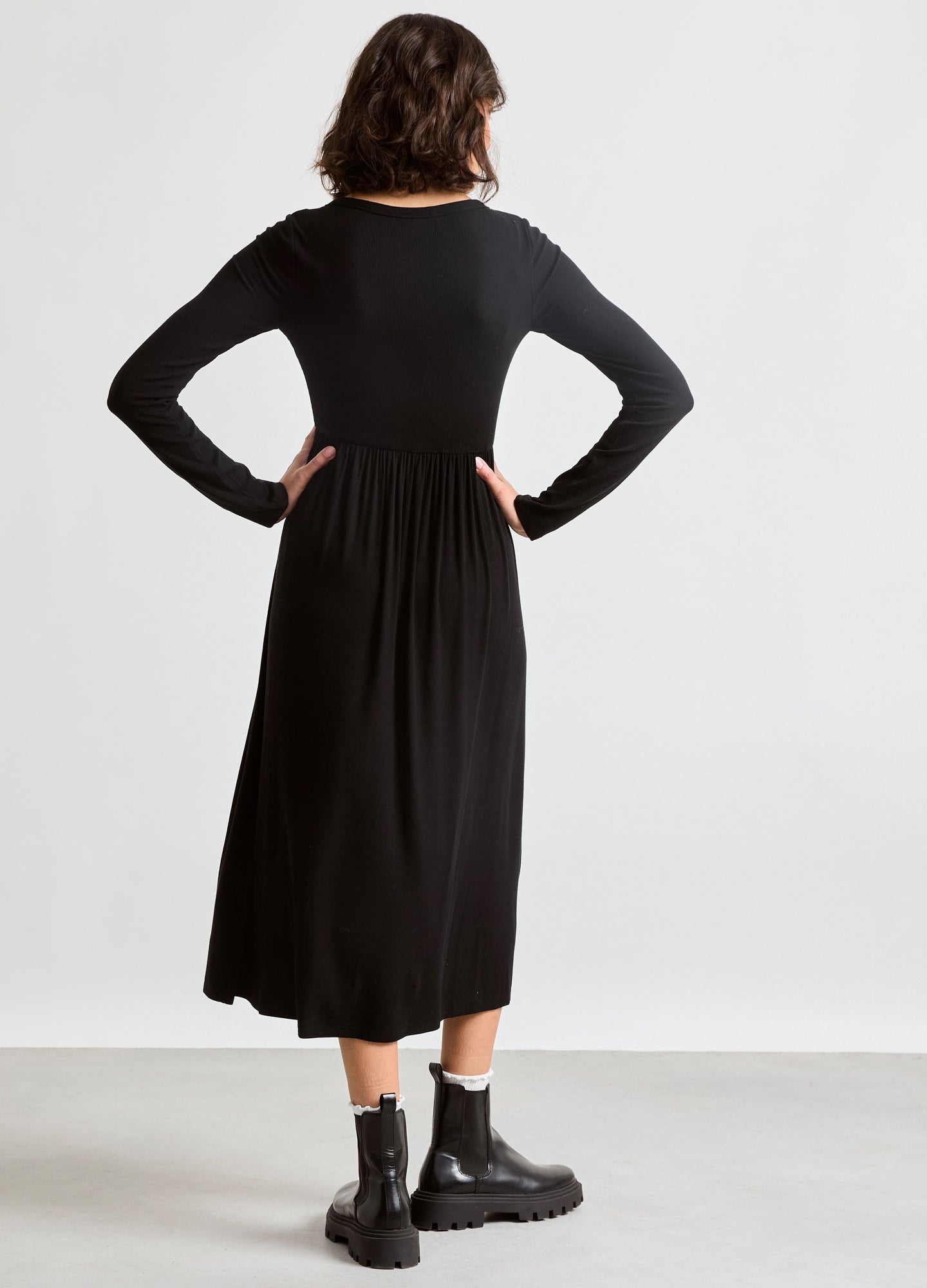 Black ribbed maternity nursing dress in midi length with long sleeves, worn by pregnant model, paired with black ankle boots.