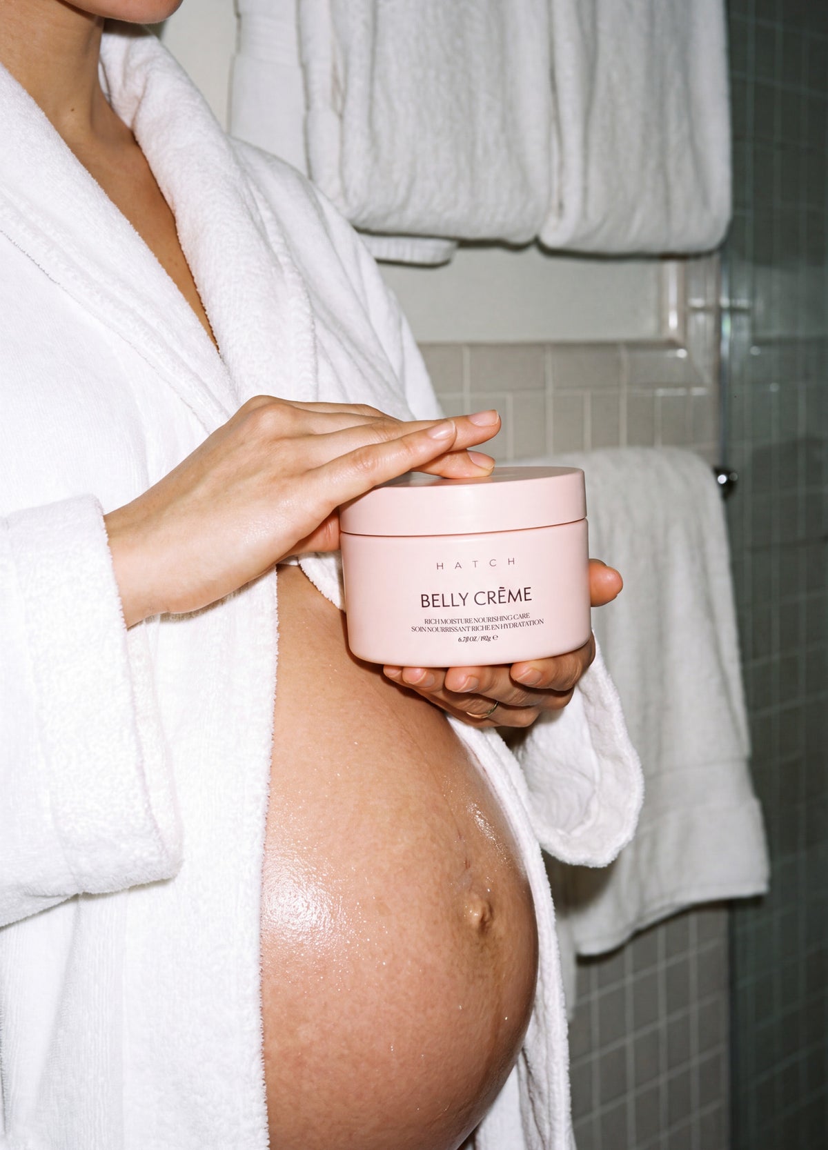 Pink round jar of Belly Crème held by a pregnant woman wearing a white robe in a bathroom setting.