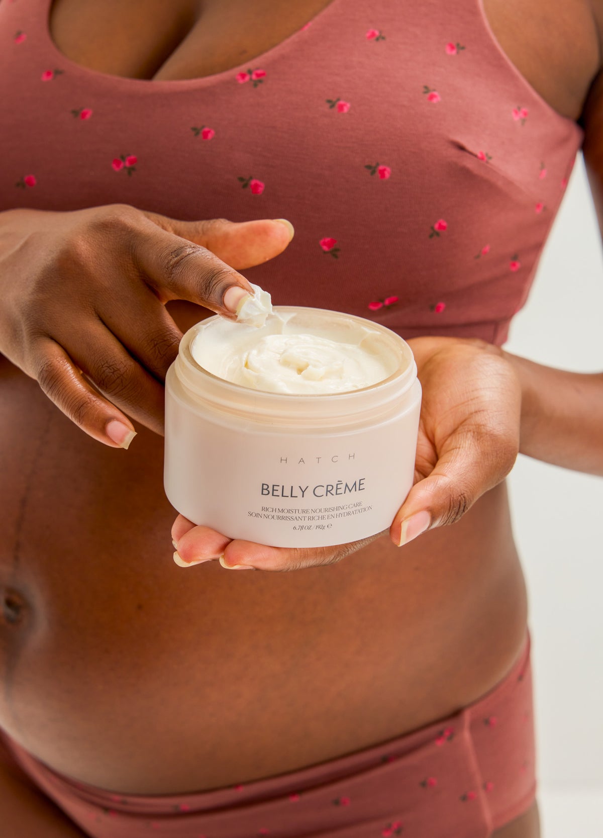 Open jar of white creamy belly moisturizer held by pregnant woman wearing mauve maternity underwear with small red heart.