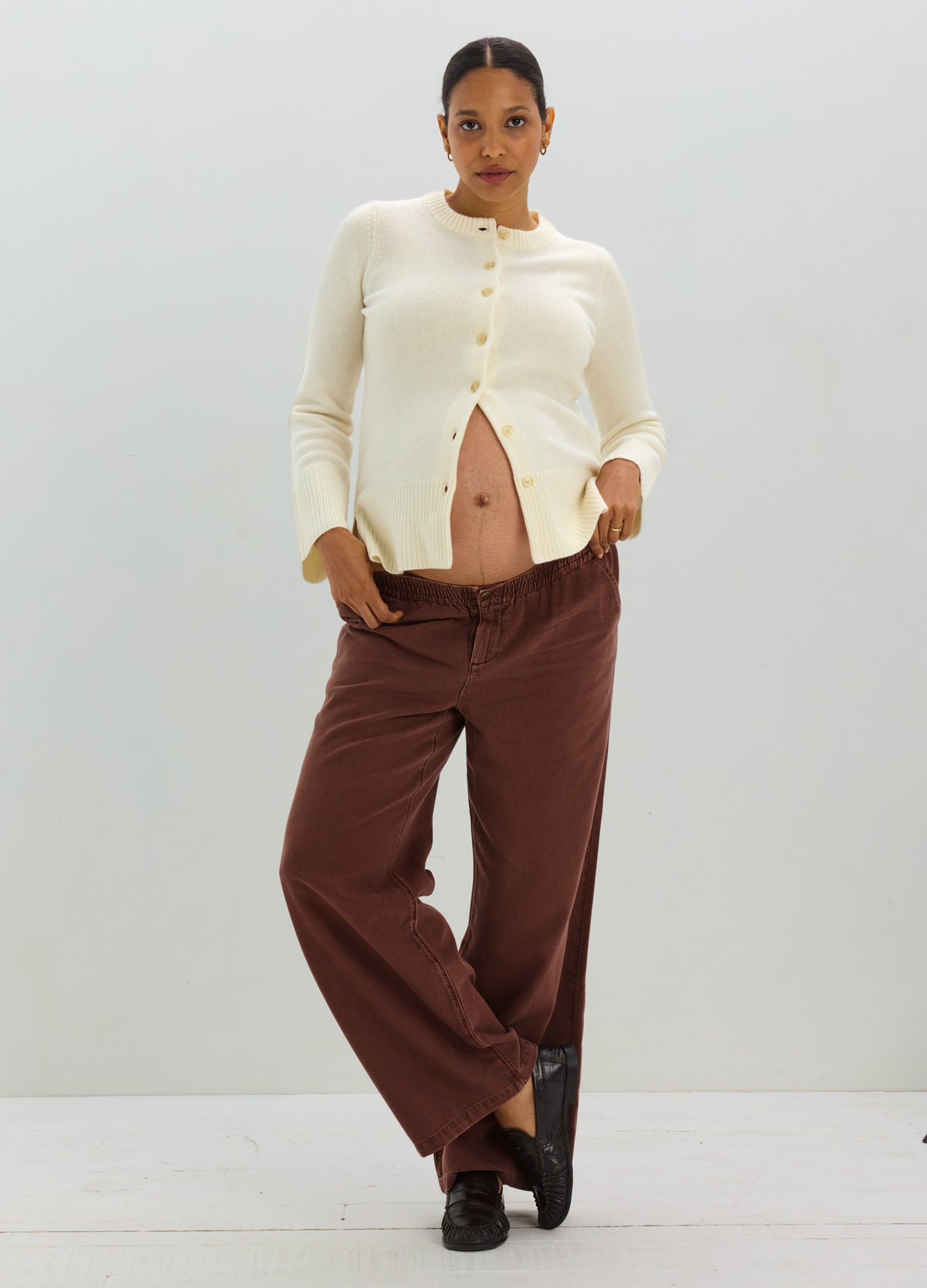 Cream-colored ribbed maternity cardigan with buttons, worn open over baby bump with brown wide-leg pants.