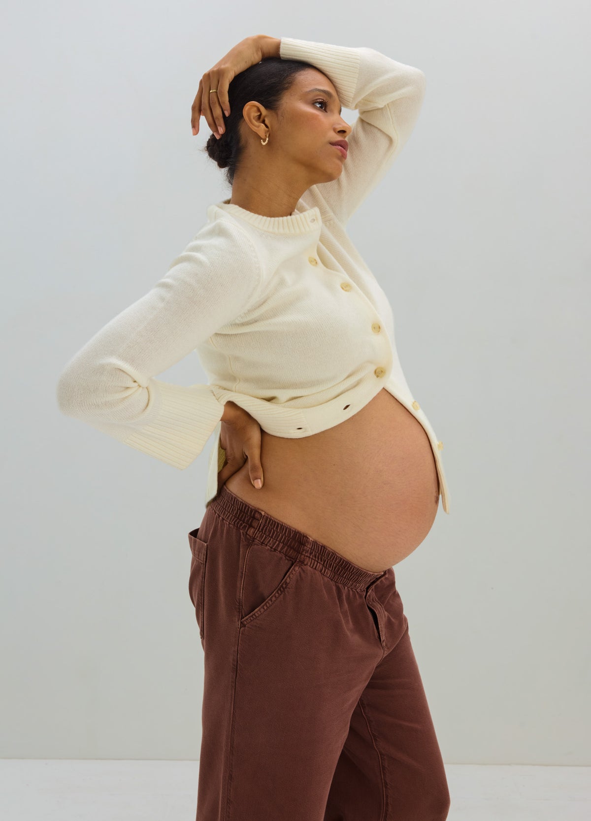 Cream-colored maternity cardigan with long sleeves and buttons, worn open over a pregnant belly, paired with loose brown.