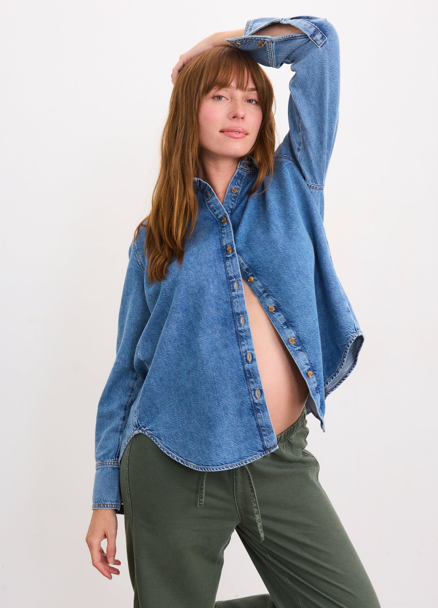 Loose-fitting blue denim shirt with buttons, styled open to reveal pregnant belly, paired with olive green pants.