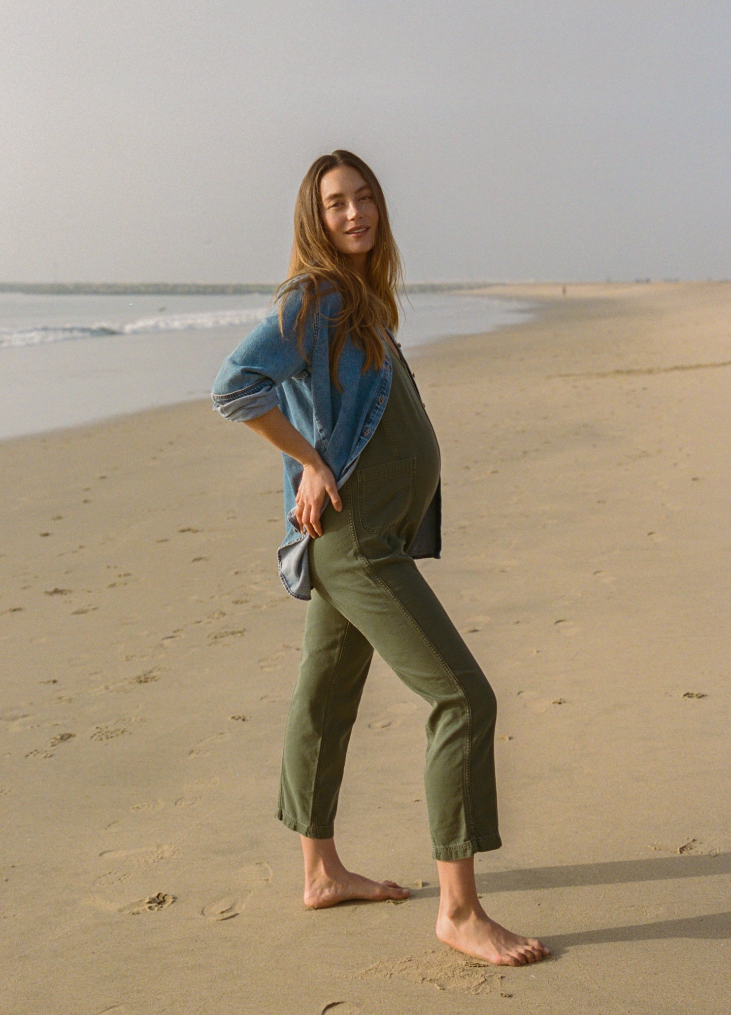 Maternity olive green drapey denim jumpsuit with side pockets, worn with loose blue shirt on beach.