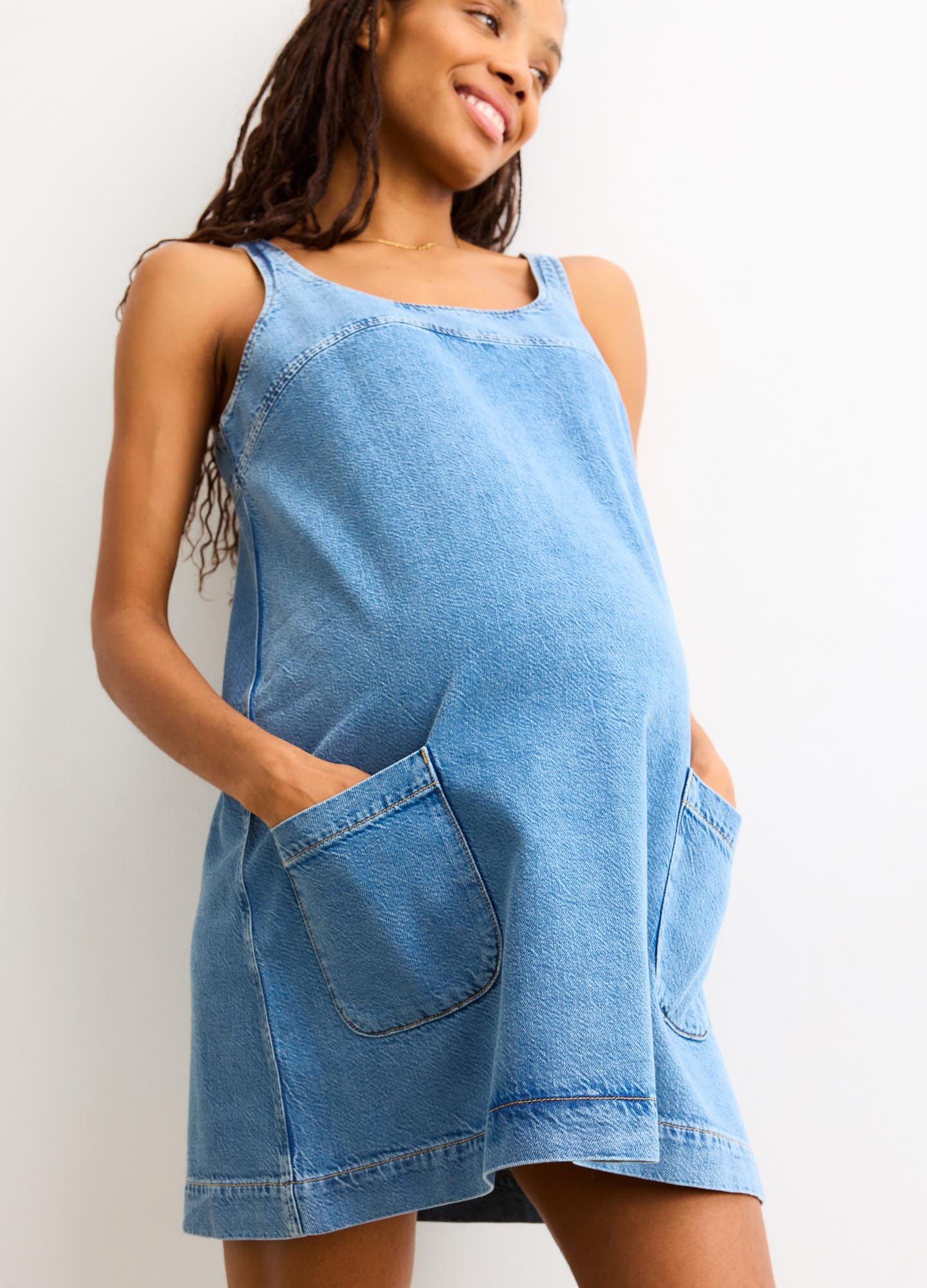 Light blue denim maternity mini dress with wide shoulder straps and front patch pockets.
