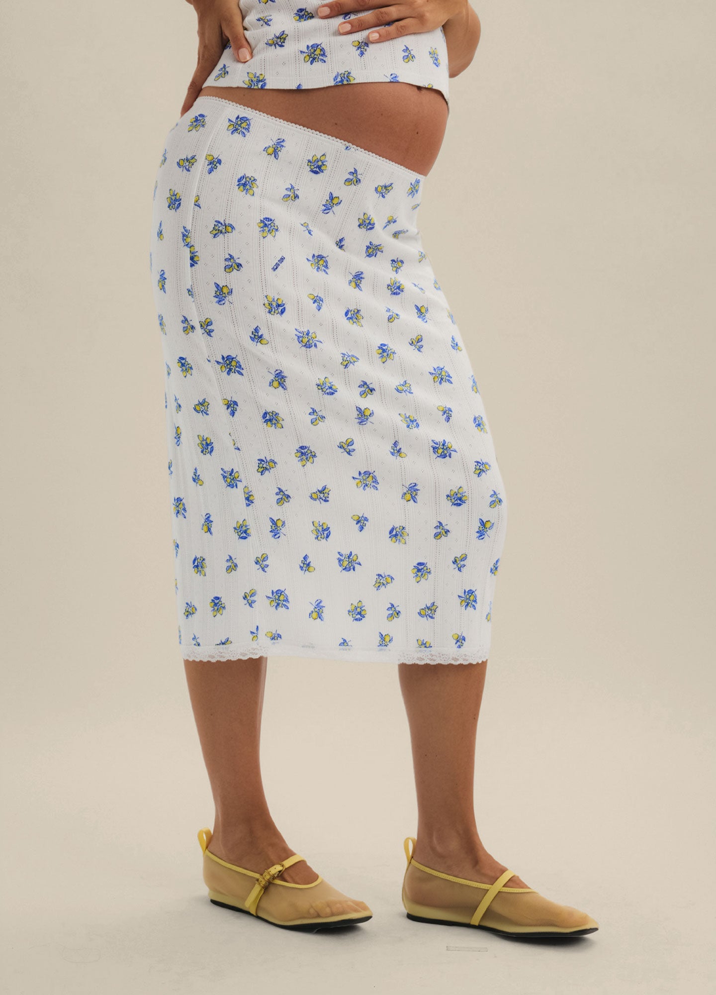 White midi maternity skirt with blue floral pointelle knit, fitted silhouette, worn with yellow sandals.