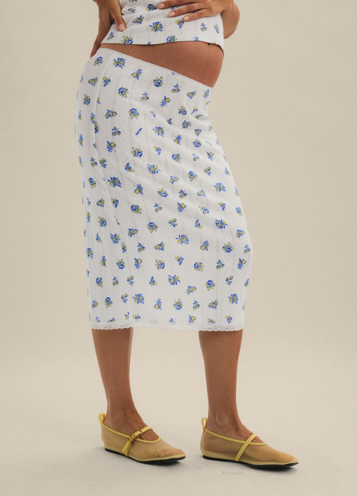 White midi maternity skirt with blue floral pointelle knit, fitted silhouette, worn with yellow sandals.