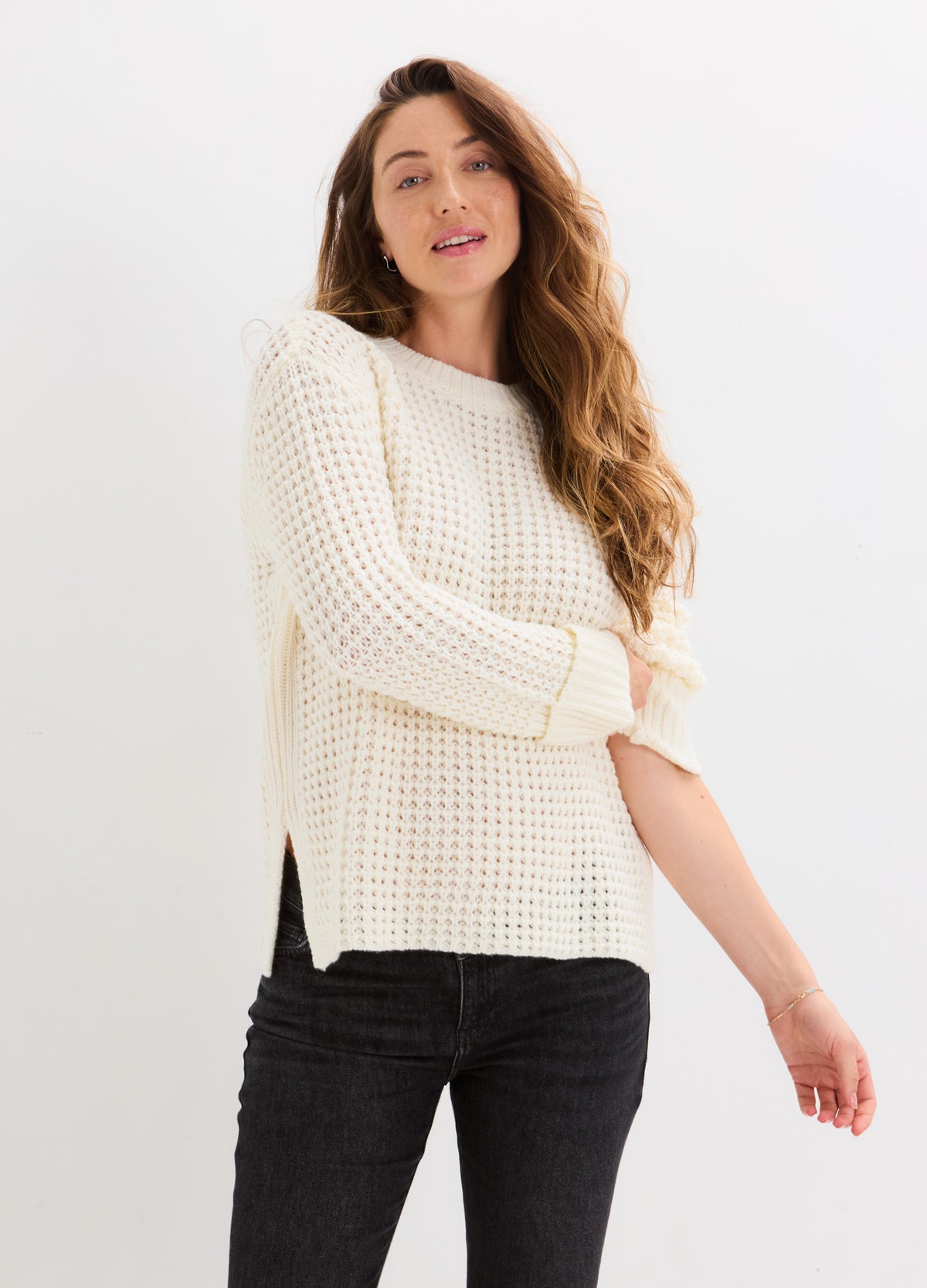 Cream waffle-knit sweater with a relaxed fit, long sleeves, and side slits, styled with black jeans.