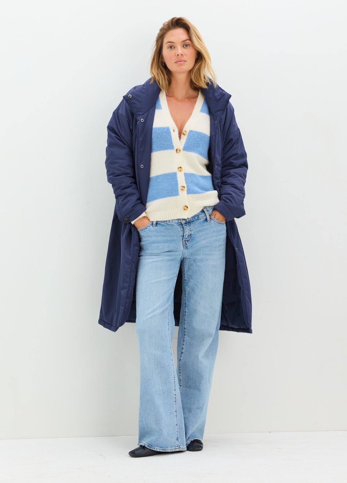 Long navy quilted coat with a relaxed fit and snap-front closure, styled over jeans and a striped sweater.