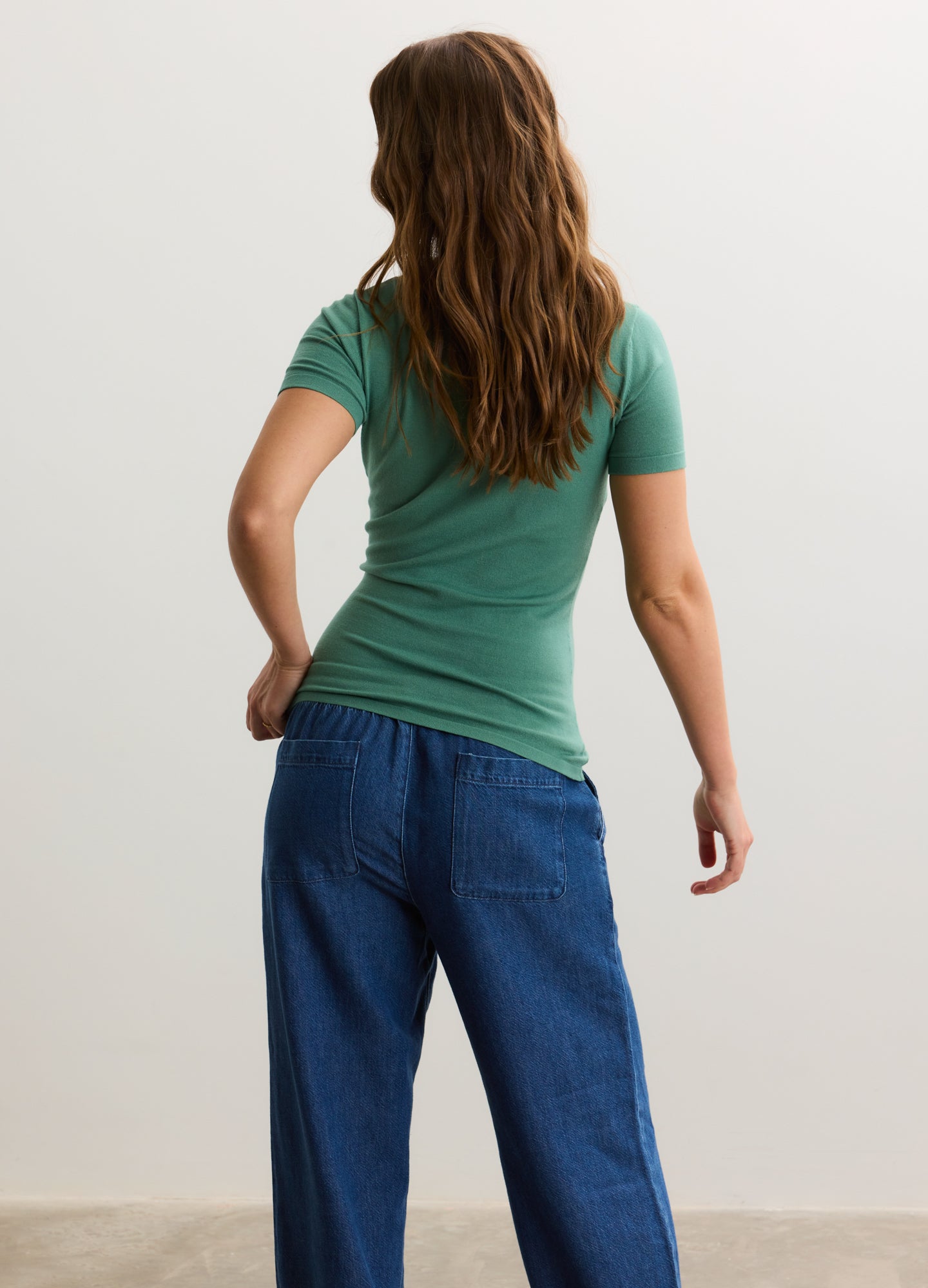 Green short-sleeve knit tee paired with loose blue maternity jeans on a pregnant model, back view.